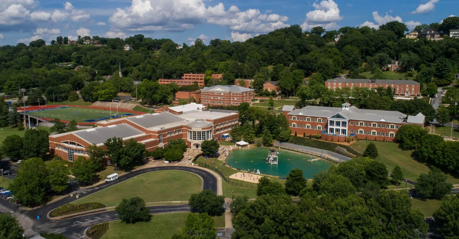 Vista aerea del campus della McCallie School con edifici in mattoni, un campo sportivo, un lago e colline alberate.