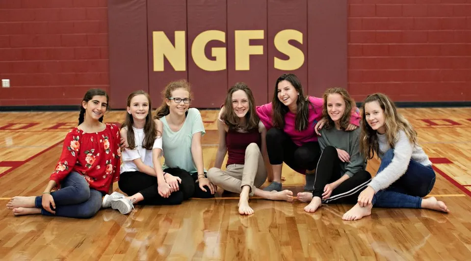 Un gruppo di studentesse siede sul pavimento della palestra della New Garden Friends School davanti alla parete con il logo NGFS.