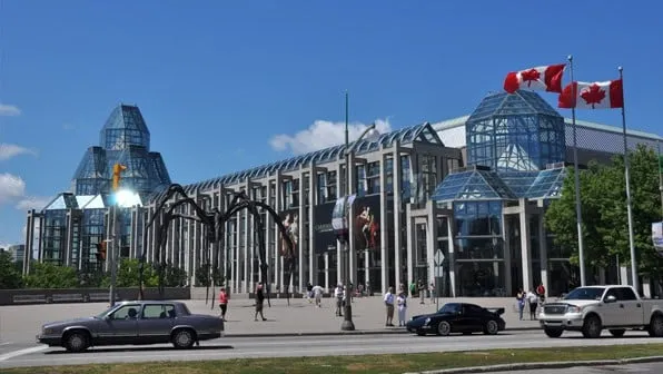 Edificio de cristal con banderas de Canadá, coches delante y una escultura gigante de araña