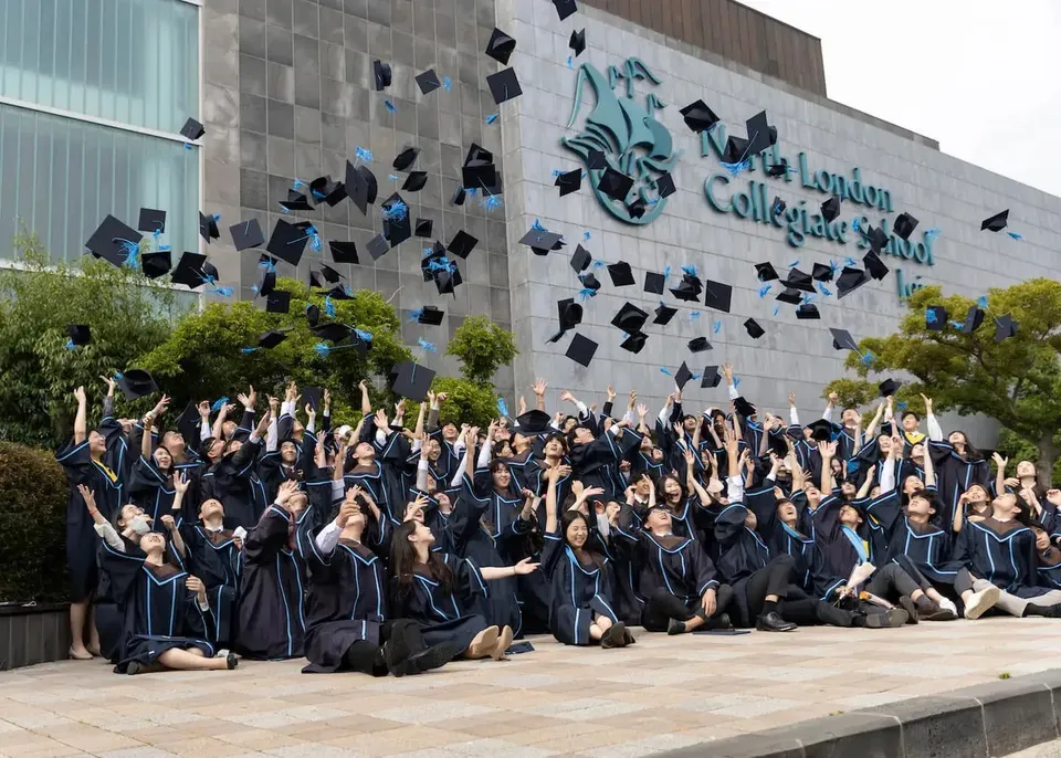 Absolventinnen und Absolventen der North London Collegiate School Jeju versammeln sich vor einem Gebäude mit einer Wandmalerei des Schullogos.