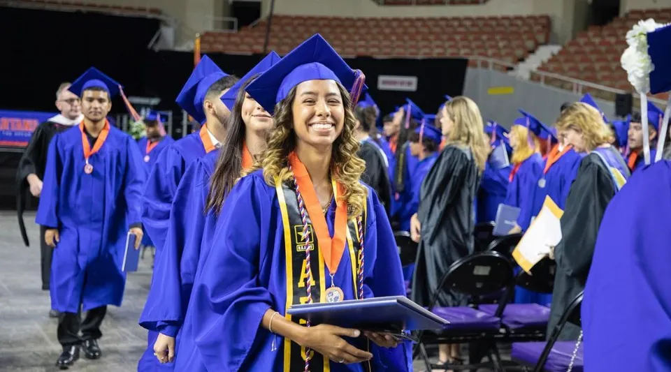 Un gruppo di diplomati del Phoenix Union High School District in piedi durante una cerimonia di diploma al chiuso, con toghe blu e diplomi in mano.