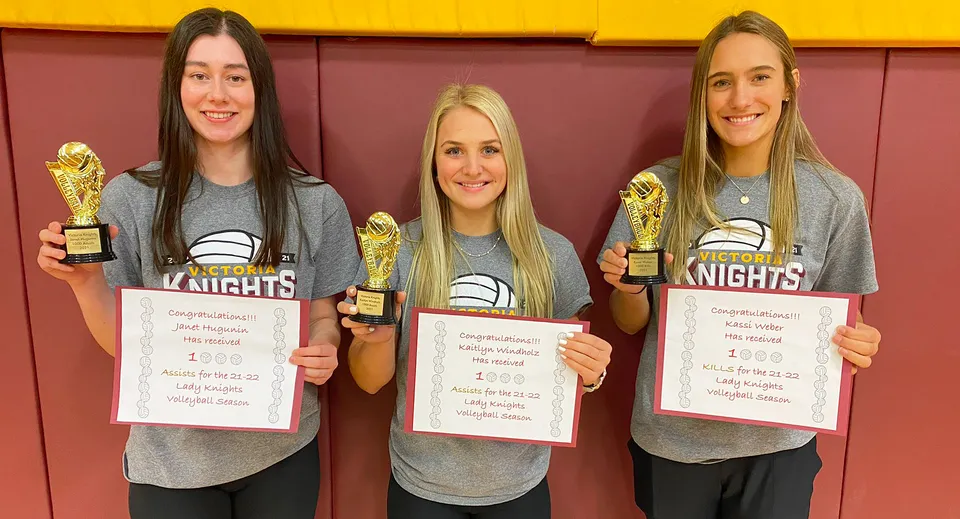 Tre studentesse della Victoria High School tengono coppe e certificati davanti a uno sfondo rosso e giallo.