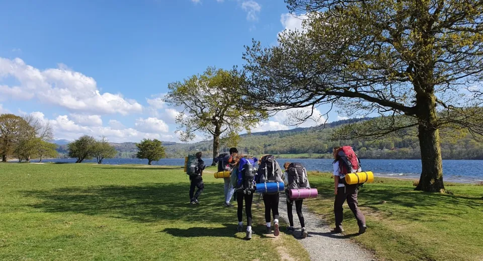 Studenti della Windermere School camminano su un prato vicino a uno specchio d'acqua tra gli alberi e sotto un cielo blu.