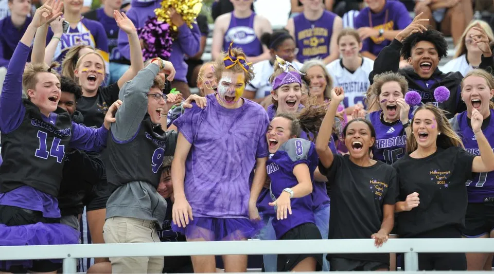 Un grande gruppo di tifosi in abiti viola e neri festeggia a un evento sportivo della Norfolk Christian Schools.