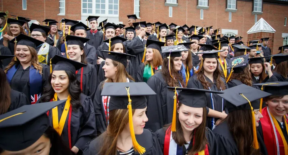 Un grande gruppo di diplomati in abito accademico festeggia davanti all'edificio in mattoni nel campus della St. Michaels University School.