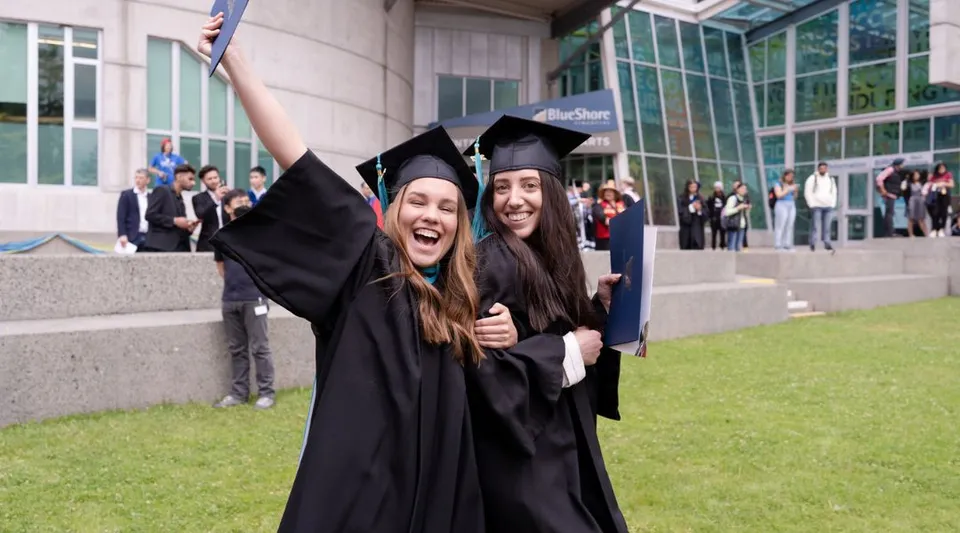Due neolaureate della Capilano University si abbracciano davanti a un edificio universitario con altri laureati sullo sfondo.