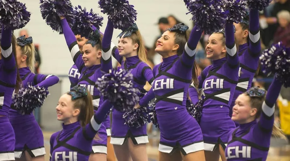 Studenti e studentesse dell'Alpine School District come cheerleader in divise viola con pompon davanti a uno striscione con la scritta EHI.