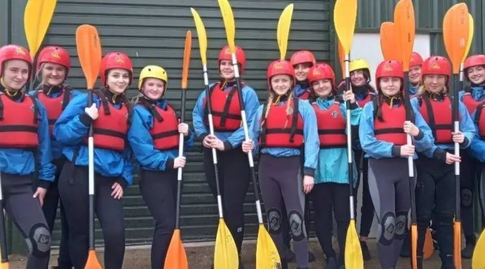 Studenti del St. Patricks College a Cork sono in piedi davanti a un edificio in metallo e tengono in mano pagaie colorate durante l'allenamento di kayak.