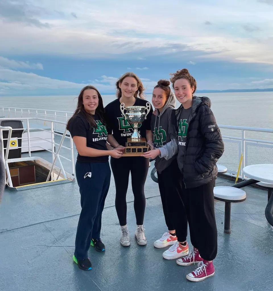 Un gruppo di quattro studenti della Nanaimo District Secondary School tiene un trofeo sul ponte di una barca con l'acqua e il cielo nuvoloso sullo sfondo.