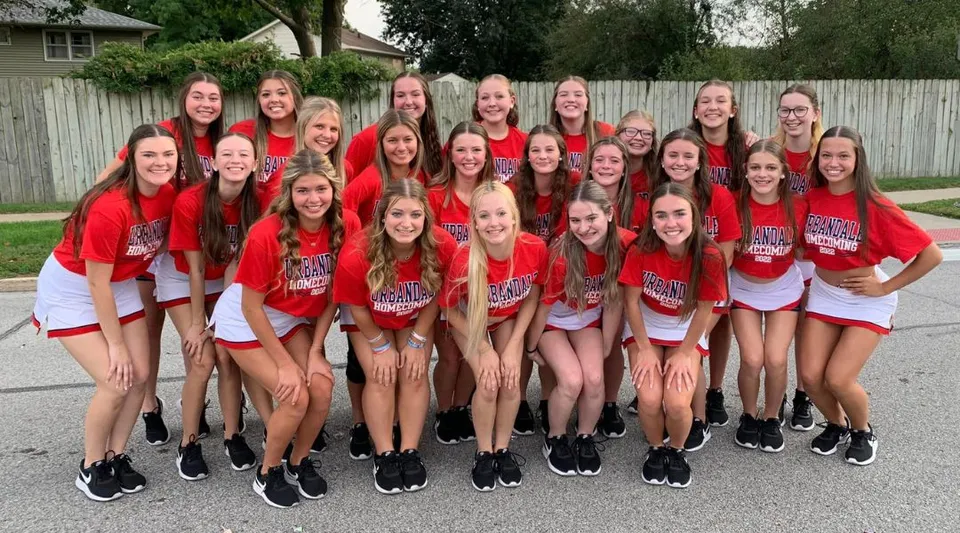 Un gruppo di studentesse in uniformi rosse è in piedi all'aperto davanti a una staccionata di legno nel campus del Urbandale Community School District.