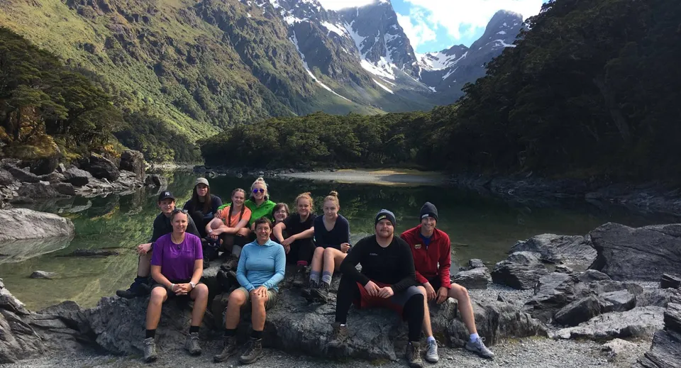 Un gruppo di studenti è seduto su rocce in un paesaggio montano con un lago e una verde vegetazione presso la Dunstan High School.