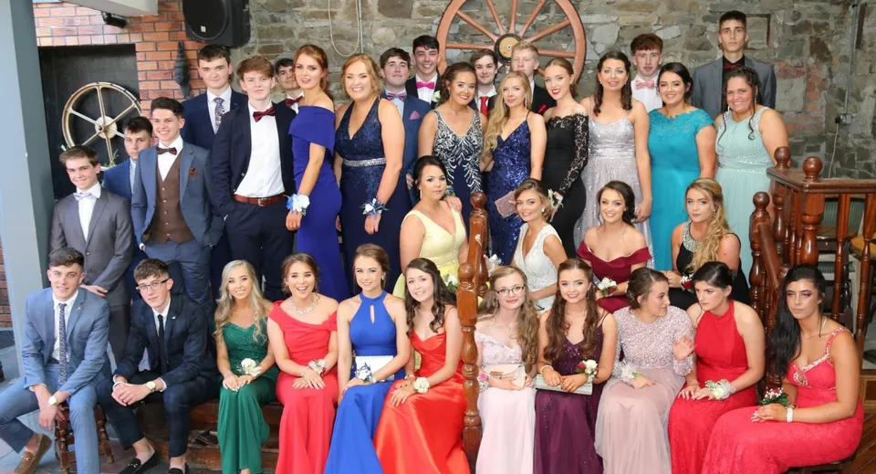 Un gruppo di studenti vestiti in modo formale nel campus della Scoil Mhuire Kanturk davanti a uno sfondo rustico con una ruota di legno.
