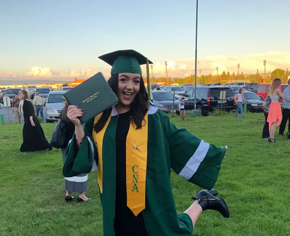 Una diplomata della Bonneville High School indossa l'abito da diploma verde e tiene un diploma davanti a un parcheggio.