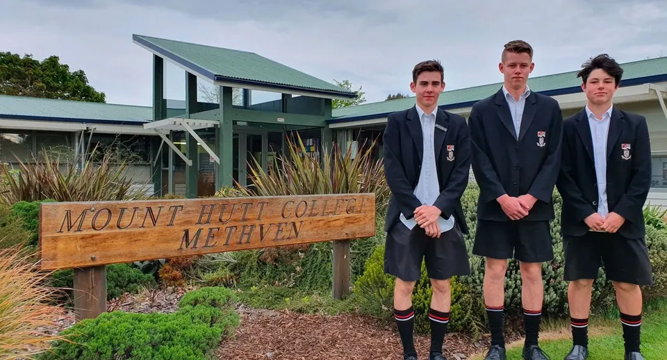 L'insegna in legno del Mount Hutt College è in primo piano, sullo sfondo tre studenti si trovano davanti a un edificio con il tetto verde.