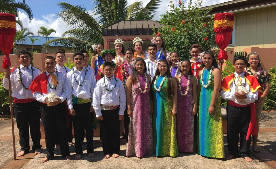Un gruppo di studenti della St. Anthony School indossa abiti tradizionali davanti a uno scenario tropicale con palme.