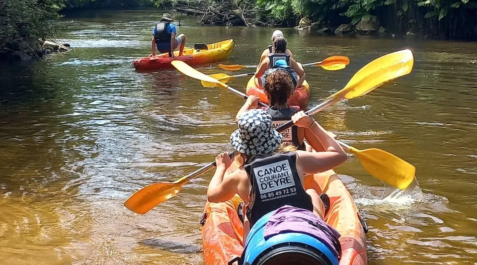 Studenti del Lycée St Elme pagaiano in kayak colorati su un fiume con una verde vegetazione spondale.
