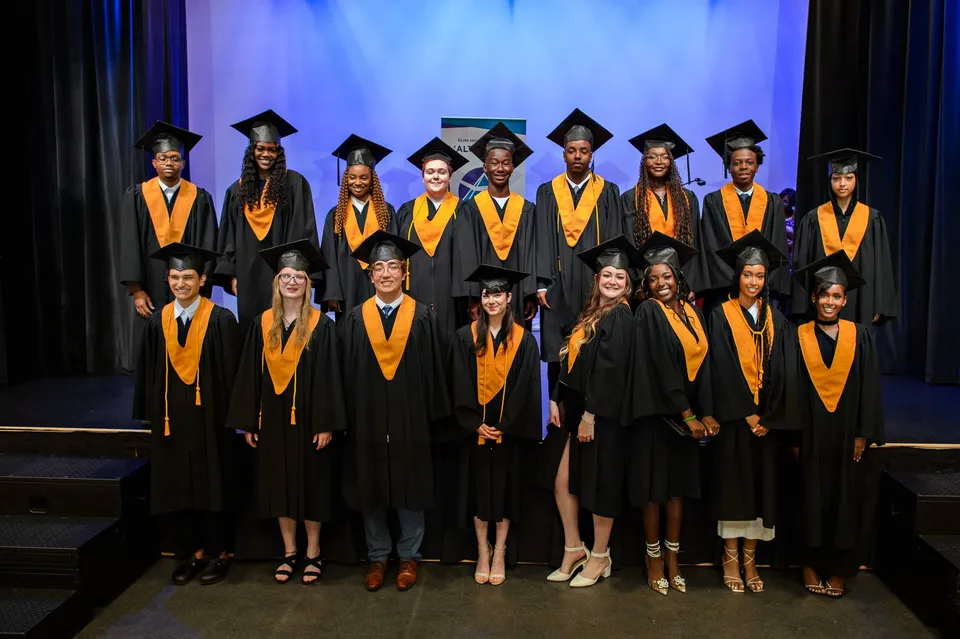 I diplomati della École secondaire publique L'Alternative sono sul palco con toghe nere e arancioni davanti a uno sfondo blu.