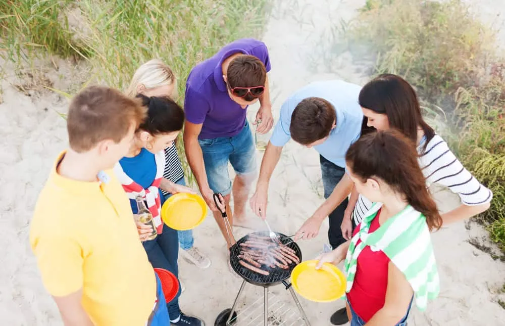 Grupo de jóvenes alrededor de una barbacoa portátil cocinando salchichas en una zona de arena