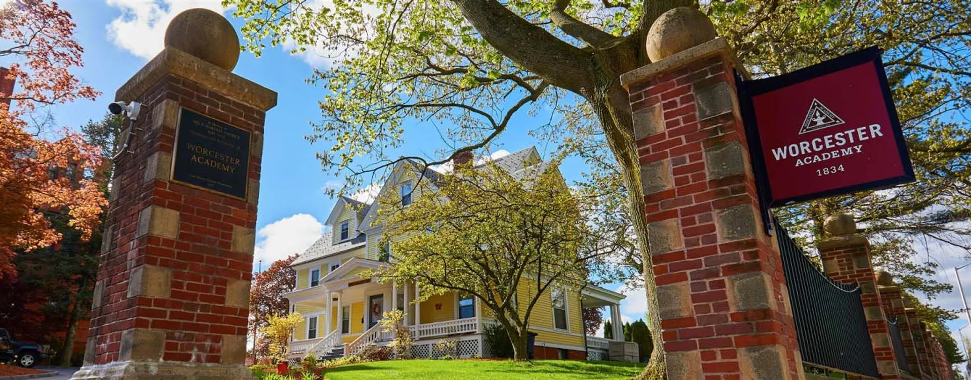 Un arco in muratura incornicia una strada alberata con una casa gialla e un edificio in mattoni rossi nel campus della Worcester Academy.