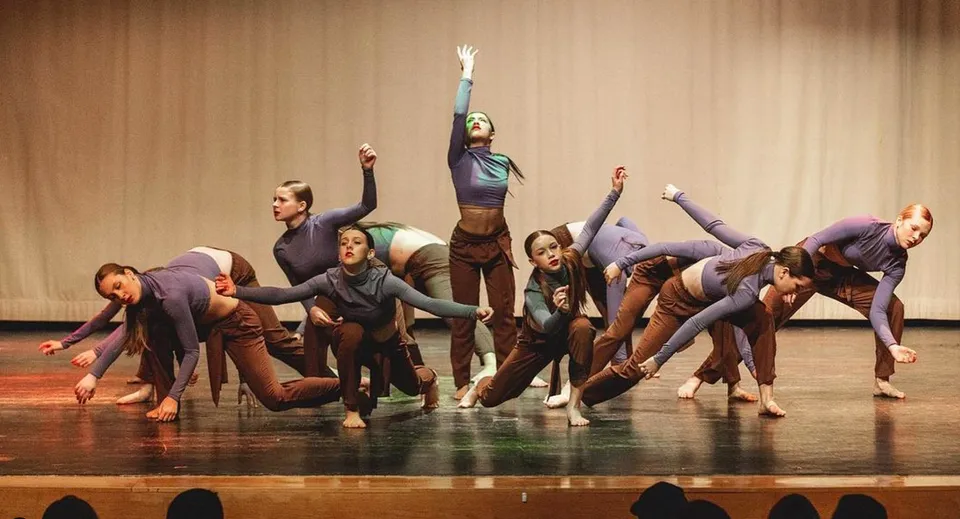 Un gruppo di ballerini si esibisce sul palco della Bear Creek Secondary School con le braccia alzate davanti a uno sfondo di legno.