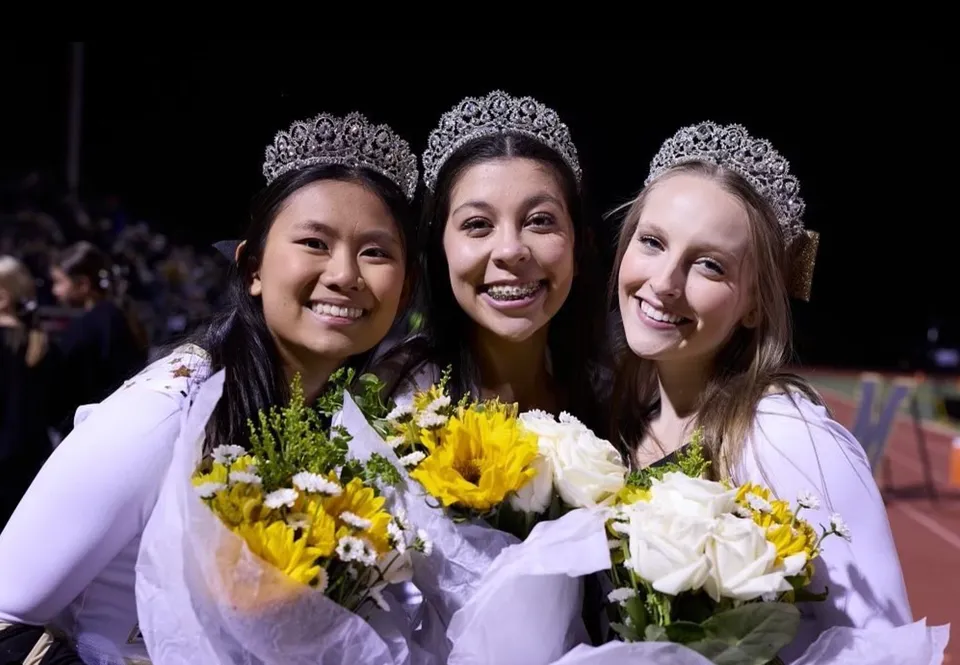 Tre studentesse della Monarch High School indossano tiare decorate e tengono in mano bouquet di fiori gialli e bianchi.