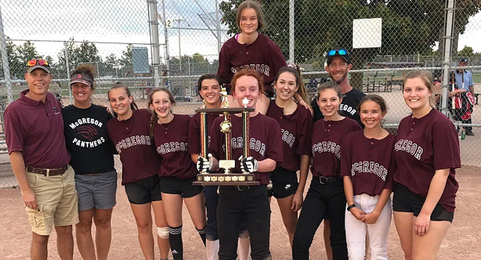 Studentesse della John McGregor Secondary School festeggiano la vittoria di un trofeo davanti a un campo sportivo.