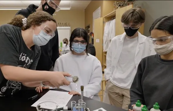 Estudiantes con mascarilla y gafas de protección realizando un experimento científico en un laboratorio escolar