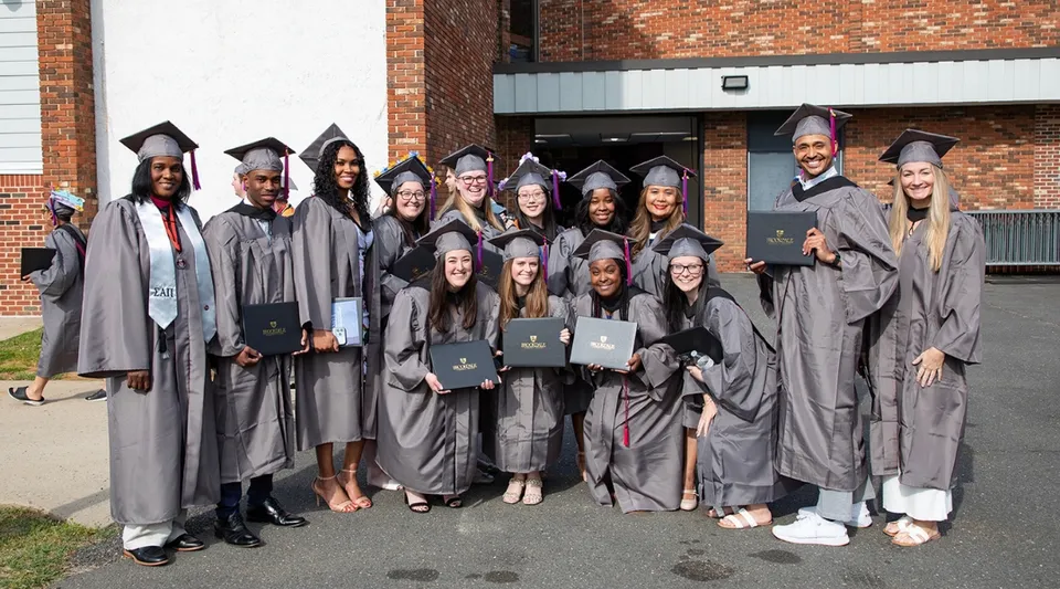 Un gruppo di diplomati in toga grigia si trova davanti a un edificio in mattoni nel campus del Brookdale Community College e mostra i propri diplomi.