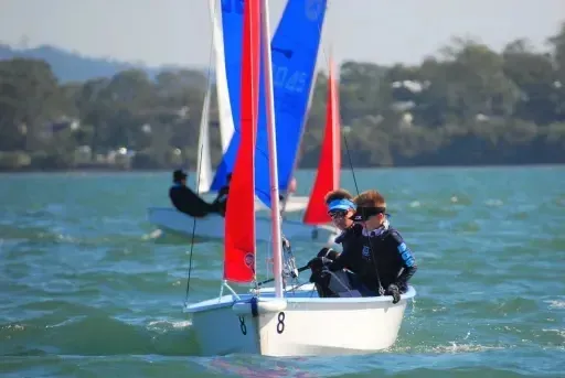 Un velero con velas de colores en un lago frente a árboles y montañas, fotografiado cerca de la Brisbane Grammar School.