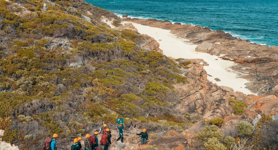 Un gruppo di studenti fa un'escursione su un sentiero costiero con vista su un oceano turchese e una spiaggia sabbiosa vicino alla Smithfield State High School.