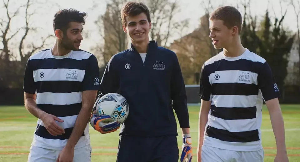 Tre studenti del DLD College London si trovano insieme su un prato con alberi e un cielo nuvoloso sullo sfondo.