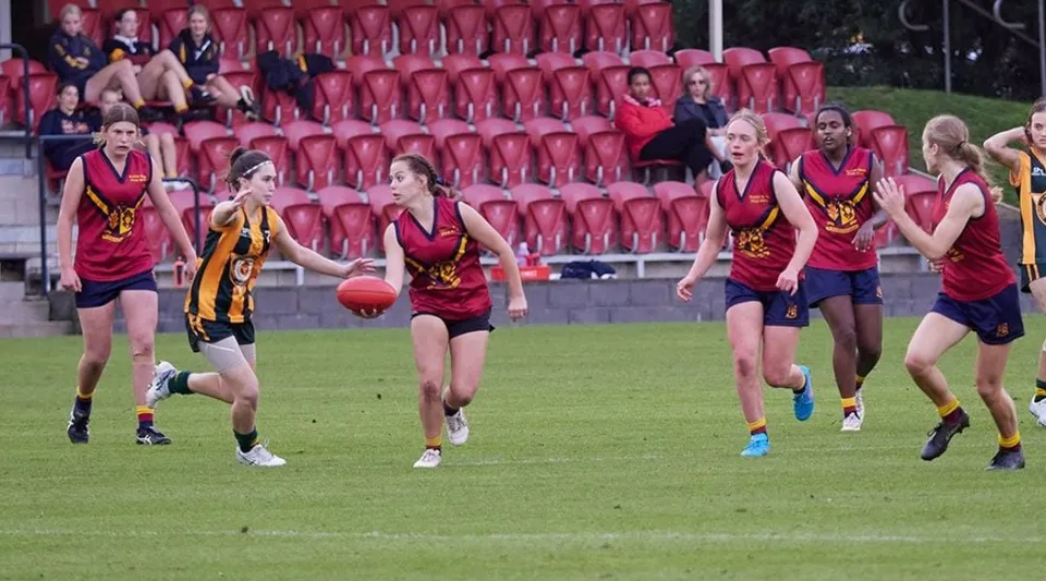 Un gruppo di giovani atlete in divisa rossa e gialla corre su un campo da gioco al Scotch Oakburn College davanti agli spettatori.
