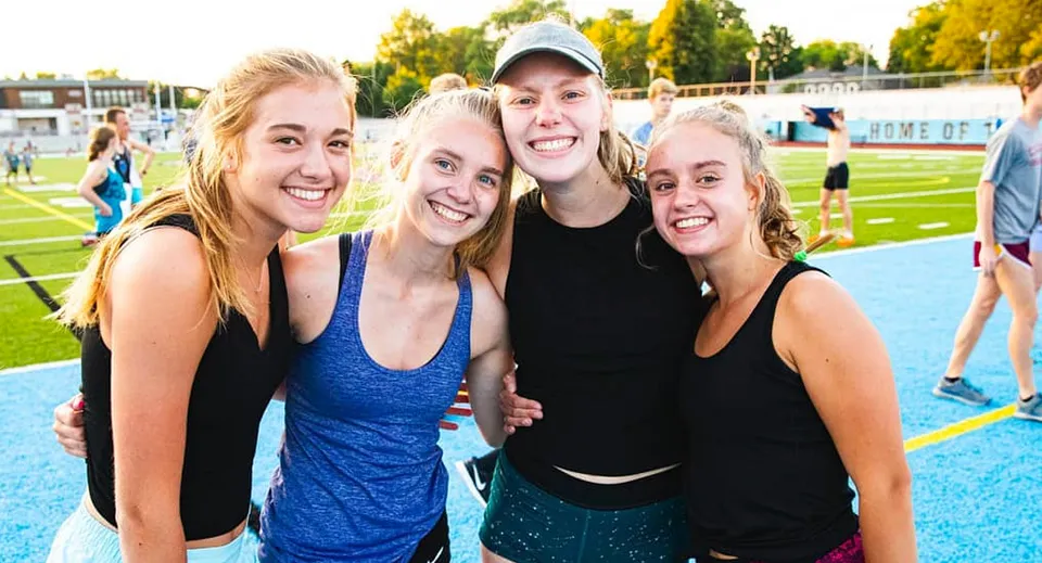 Quattro studentesse della Grand Rapids Christian High School posano sorridenti su un campo sportivo con alberi ed edifici sullo sfondo.
