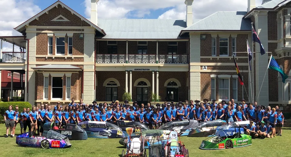 Studenti della Maryborough State High School si radunano davanti a un edificio storico accanto a diverse auto da corsa.