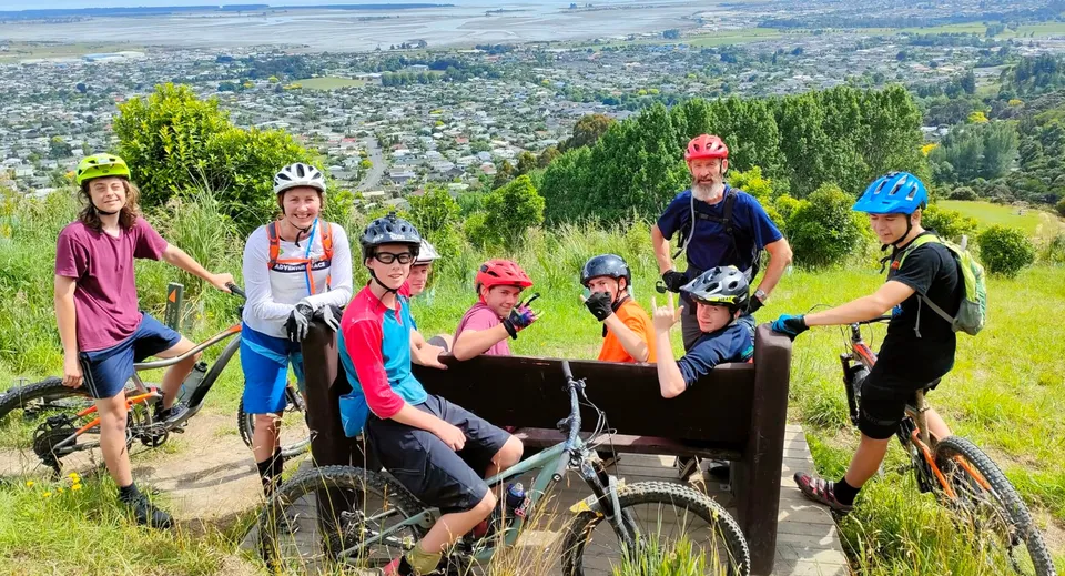 Un gruppo di studenti e studentesse della Motueka High School in abbigliamento da bici colorato posa su un sentiero di montagna con vista sulla città sullo sfondo.