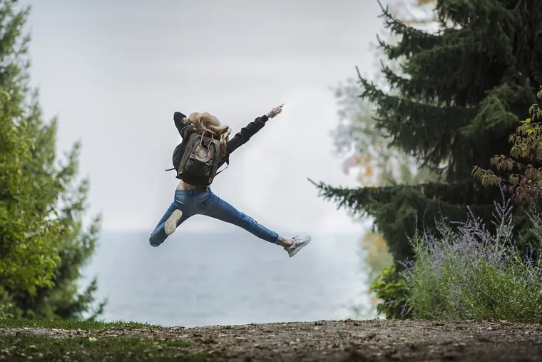 Junge Frau mit Rucksack springt mit ausgestreckten Armen und Beinen an einem Aussichtspunkt