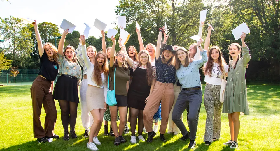 Un gruppo di giovani studentesse della Burgess Hill Girls sta con le braccia alzate su un prato all'aperto davanti agli alberi.