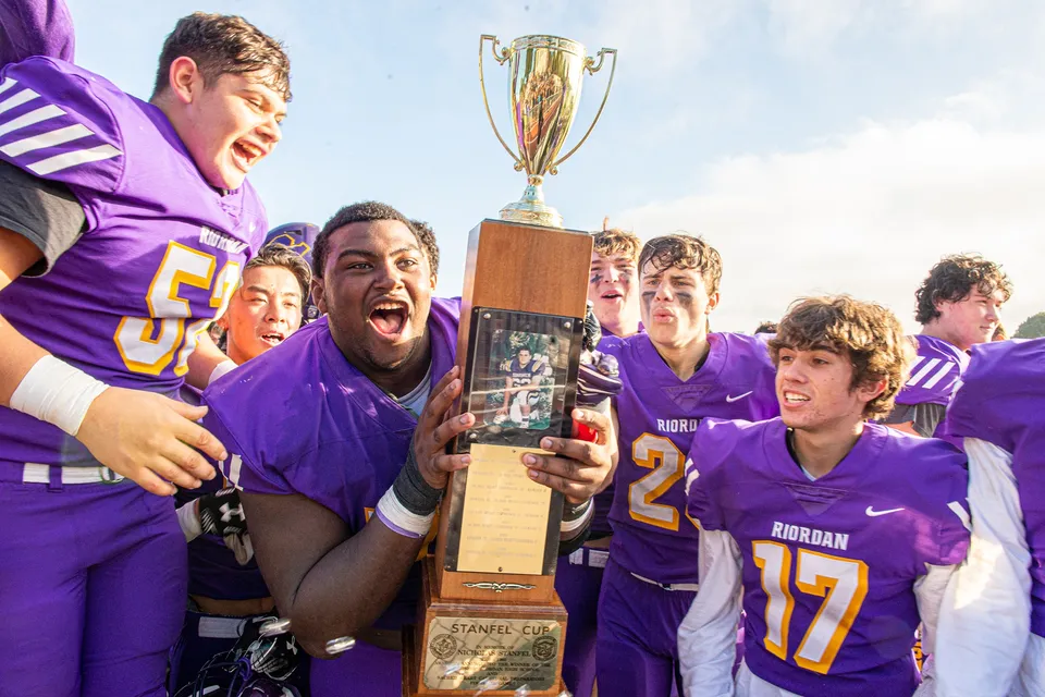 Studenti della Archbishop Riordan High School festeggiano con un trofeo davanti a uno stadio sotto un cielo blu.