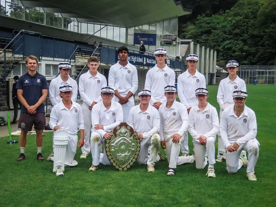 Un gruppo di studenti della Otago Boys' High School posa su un campo da cricket con uno stadio sullo sfondo.