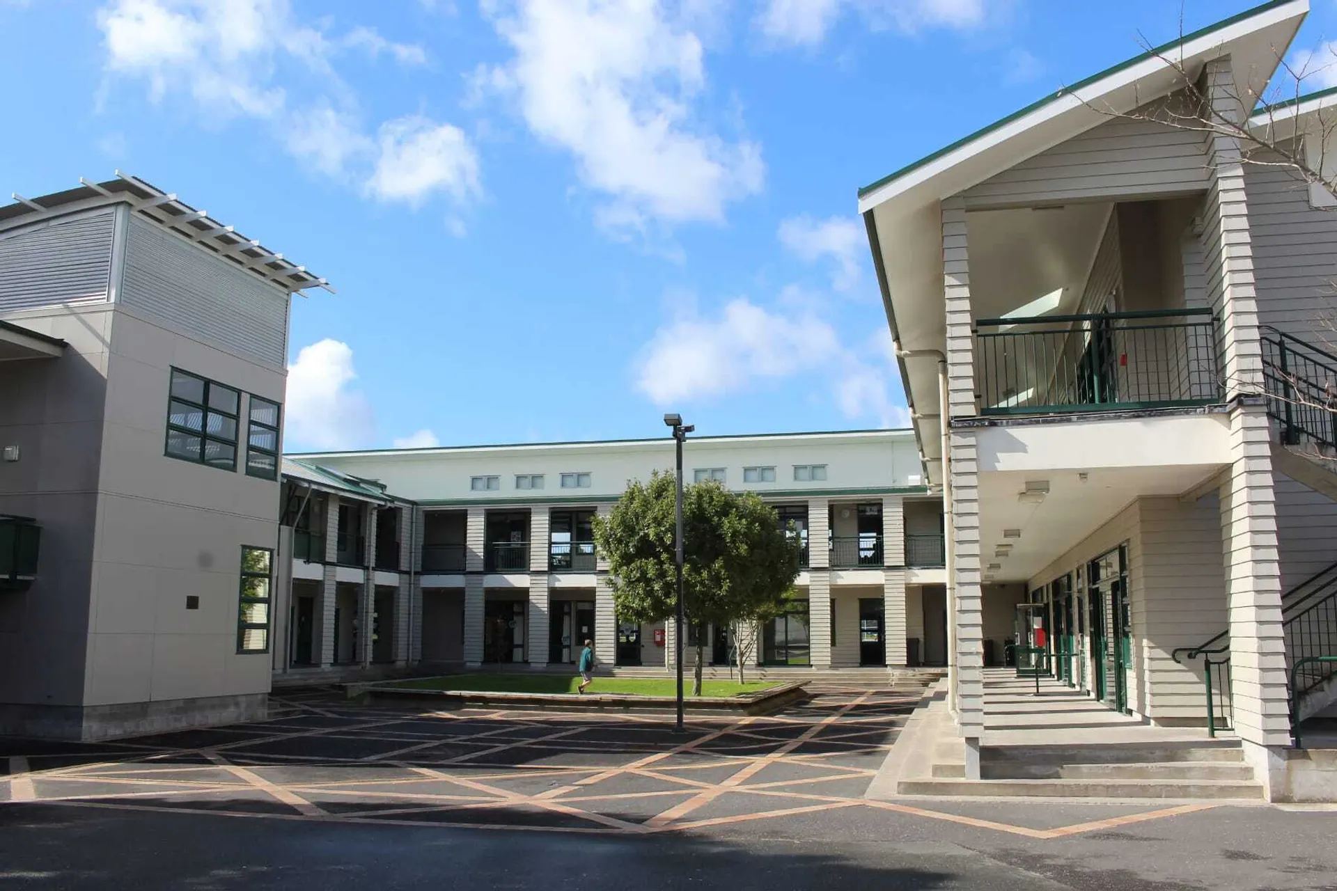 L'edificio principale a più piani dell'Orewa College con cortile interno e sentieri, circondato da alberi sotto un cielo azzurro.