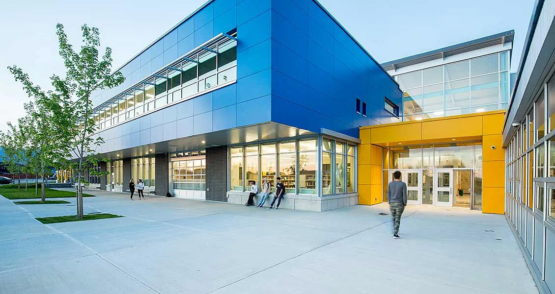 El edificio de varias plantas de Alpha School con fachada azul y detalles amarillos está rodeado por una acera y vegetación.