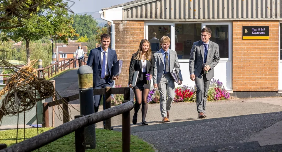 Un gruppo di persone cammina lungo un viale del campus del Queen’s College circondato dal verde e con edifici sullo sfondo.