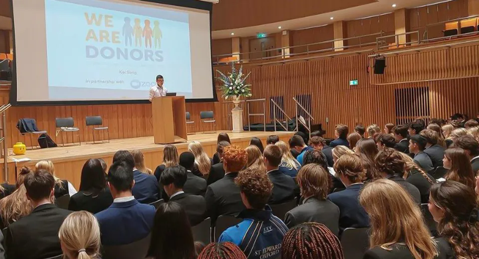 Una conferenza nel grande auditorium della St Edward's School (Oxford) con un relatore al podio e una proiezione con il testo We are donors.