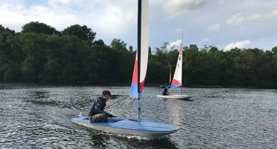 Una persona va in barca a vela con una piccola imbarcazione sull'acqua sullo sfondo di alberi nella tenuta del Clifton College.