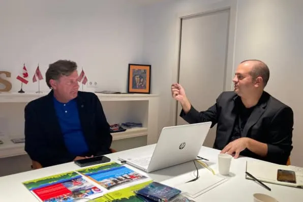 Dos hombres conversando en una oficina con un portátil y folletos sobre la mesa
