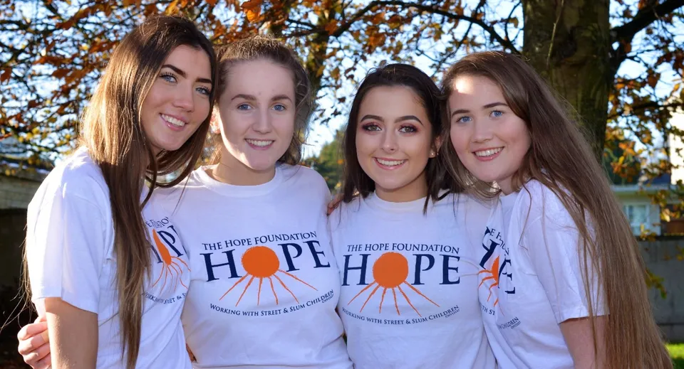 Quattro studentesse della St. Anne's Secondary School sono davanti a un albero dal fogliame autunnale e indossano t-shirt bianche con il logo HOPE.