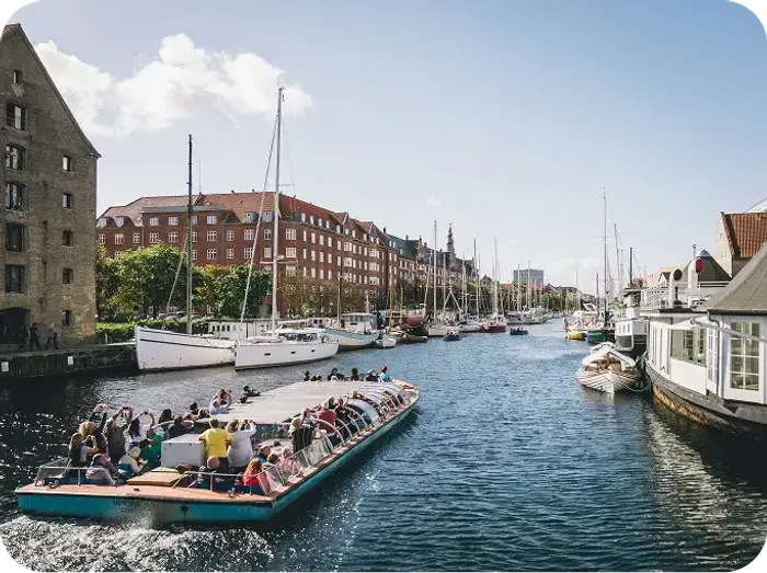 Barca turistica affollata naviga lungo un canale di Copenhagen tra edifici storici e velieri ormeggiati