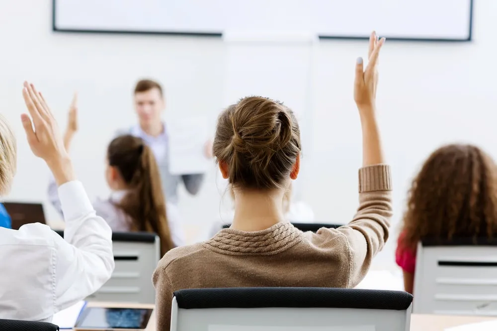 Schülerinnen und Schüler heben im Klassenzimmer die Hand, vorne steht ein junger Lehrer