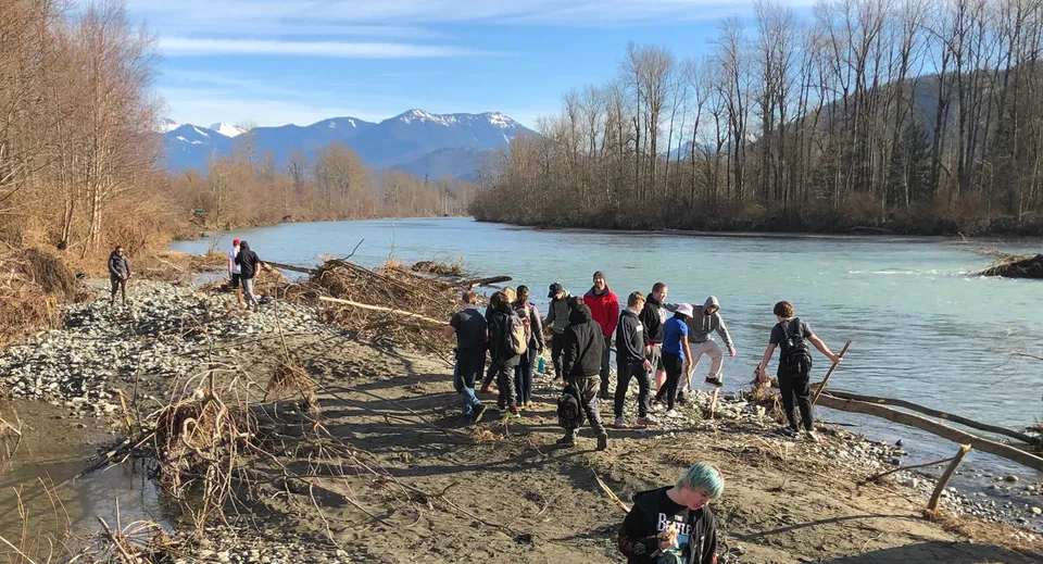 Un gruppo di studenti della Imagine High Integrated Arts and Technology Secondary School cammina lungo un fiume su un sentiero fangoso con montagne innevate sullo sfondo.