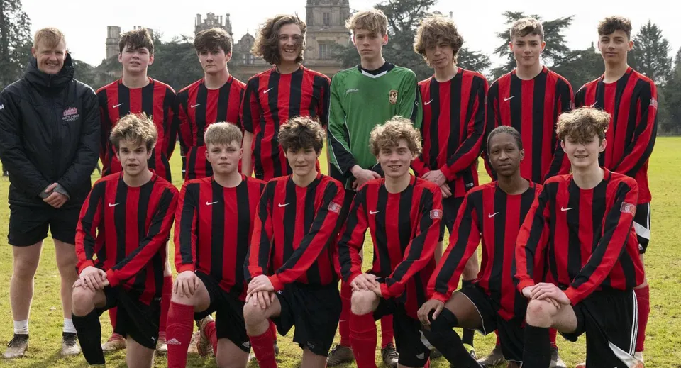 Un gruppo di giovani calciatori con maglie rosse e nere si trova sul campo in erba della Westonbirt School davanti al campanile di una chiesa.